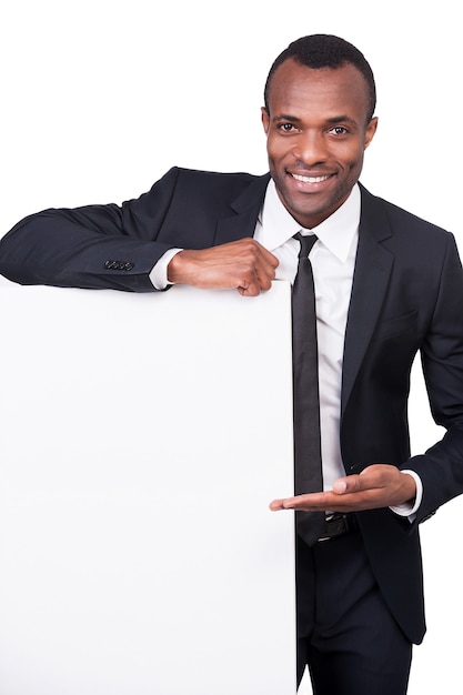 Hombre de negocios apuntando un espacio de copia. Apuesto joven africano en traje completo apuntando copia espacio y sonriendo mientras está de pie aislado sobre fondo blanco.