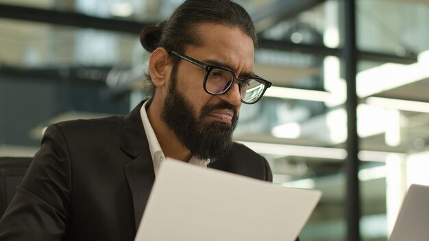 hombre de negocios árabe musulmán hombre de negocios indio empleador empresario gerente confundido con papeles y portátil en la oficina problema papeleo mal resultado hablando llamada de video trabajando con documentos fracaso