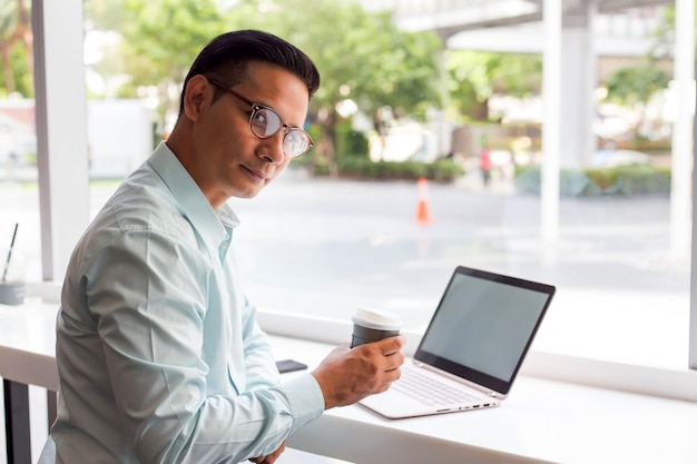 Hombre de negocios de Asia que sostiene la taza de café caliente y que trabaja con el ordenador portátil mientras que se sienta cafetería. Concepto de hombres de negocios jovenes