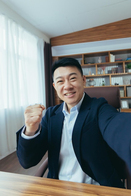 Un hombre de negocios asiático alegre y divertido mira la cámara de un teléfono inteligente habla en una videollamada un hombre trabaja en la oficina hace un gesto de felicitación