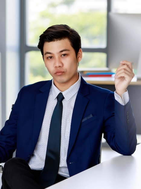 Hombre de negocios asiático atractivo joven con traje azul marino con camisa blanca y corbata sentado en la silla ejecutiva haciendo cara de confianza en la oficina con iluminación natural.