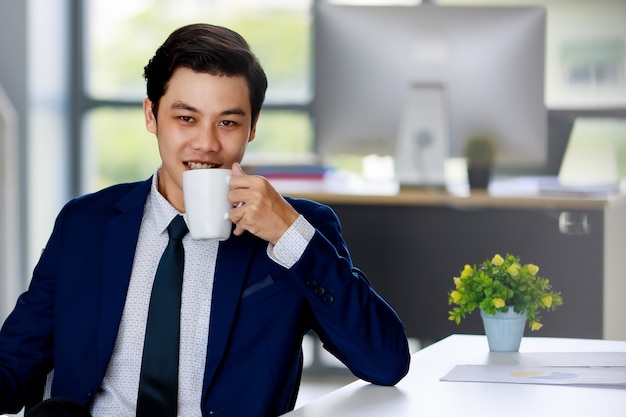 Hombre de negocios asiático atractivo joven con traje azul marino con camisa blanca y corbata sentado en la silla ejecutiva tomando café en la oficina con iluminación natural.