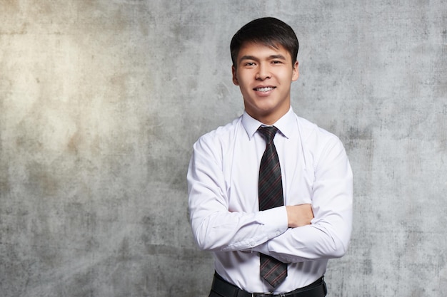 Hombre de negocios asiático en camisa y corbata sobre fondo gris. Retrato de un exitoso empresario kazajo sonriente con los brazos cruzados.