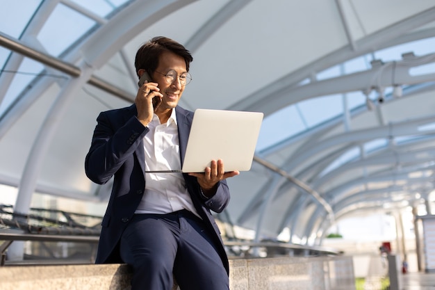 Hombre de negocios asiático con confianza en gafas y traje que trabaja en la computadora portátil y el teléfono móvil al aire libre en la ciudad.