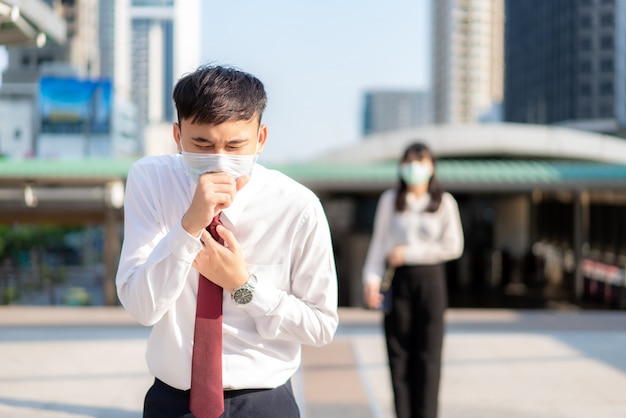 Hombre de negocios asiático enfermo tos con máscara con empresaria en segundo plano para mantener la distancia, proteger contra los virus COVID-19 y el distanciamiento social de las personas por riesgo de infección