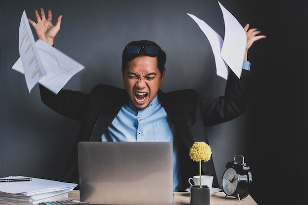 Un hombre de negocios asiático enojado tira un informe de negocios mientras grita en el escritorio de la oficina aislado en gris