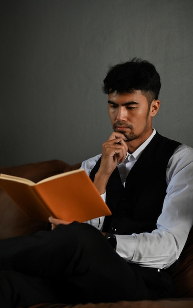 Hombre de negocios asiático exitoso e inteligente en traje formal sentado en el sofá y leyendo un libro