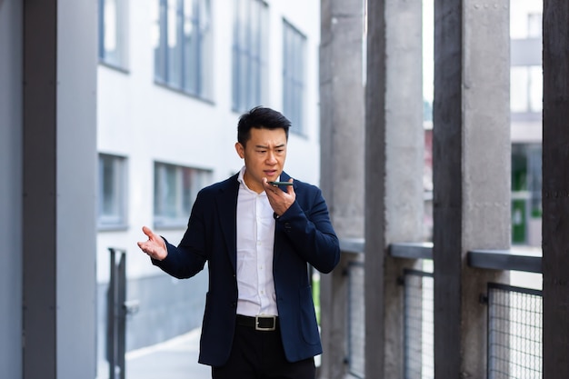 Hombre de negocios asiático exitoso enojado y serio explica información a los empleados que usan el teléfono, habla cerca de la oficina afuera