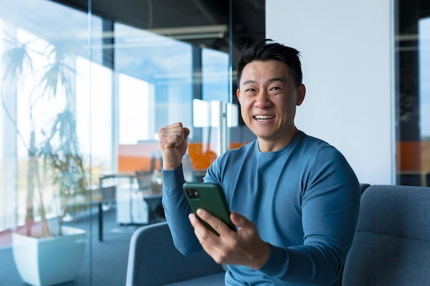 Hombre de negocios asiático exitoso y feliz celebrando la victoria sentado y trabajando en la oficina moderna en el escritorio celebrando la victoria mirando a la cámara y gritando alegremente sosteniendo el teléfono móvil