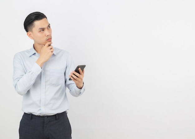 Hombre de negocios asiático guapo joven sosteniendo un teléfono inteligente negro y pensando en una idea mientras mira hacia arriba