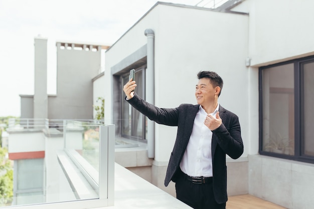 Hombre de negocios asiático hablando por teléfono de vídeo de pie en el balcón terraza
