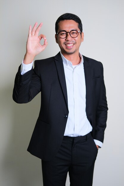 Hombre de negocios asiático joven sonriendo feliz mientras da señal de dedo OK