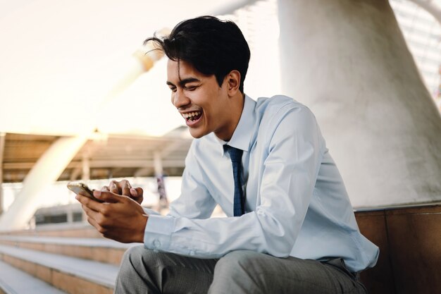 Hombre de negocios asiático joven sonriente que usa el teléfono móvil en la ciudad. Sentado en la escalera