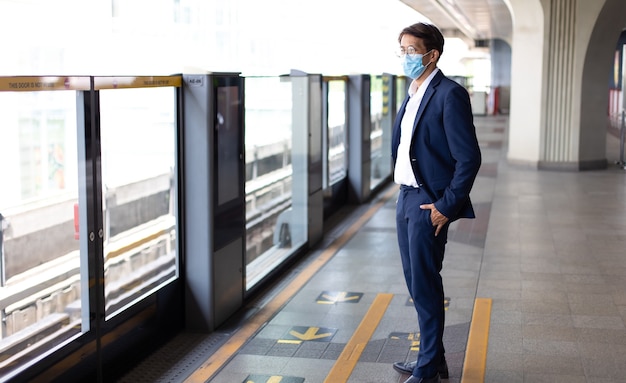 Hombre de negocios asiático con máscaras faciales mientras viaja en la estación de transporte público durante la pandemia de COVID-19.