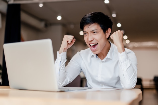 Hombre de negocios asiático milenario en la oficina con laptop feliz con su victoria