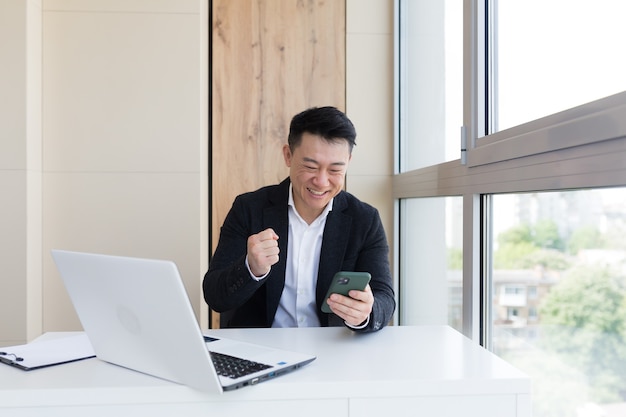 Hombre de negocios asiático en la oficina mirando el teléfono móvil con emoción ganador o ganar