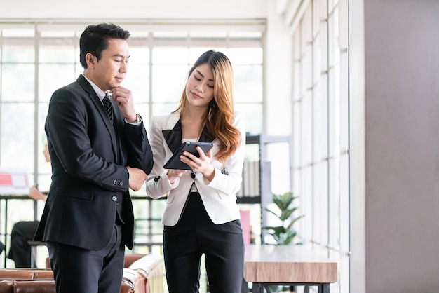 Un hombre de negocios asiático senior y una joven mujer de negocios discuten en la oficina trabajando en la estrategia de las redes sociales mirando una tableta con colegas borrosos en la imagen de fondo con espacio de copia