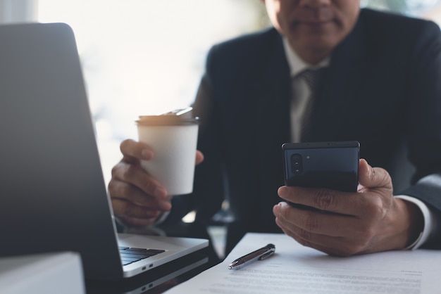 Hombre de negocios asiático mediante teléfono móvil y tomando café trabajando en la oficina