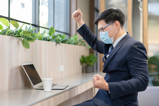 Hombre de negocios asiático en traje y con máscara médica protectora levantando su brazo para celebrar el logro del éxito laboral mientras mira la computadora portátil en el lugar de trabajo