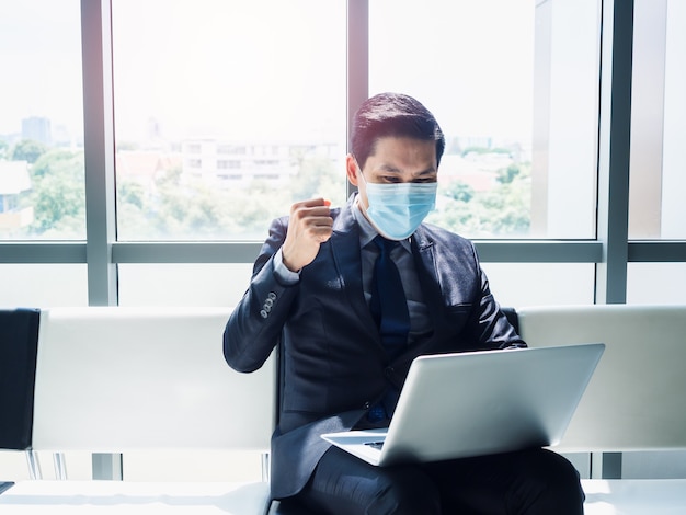 Hombre de negocios asiático en traje con mascarilla protectora levantó la mano con alegría y alegría cuando vio un monitor de computadora portátil en su regazo