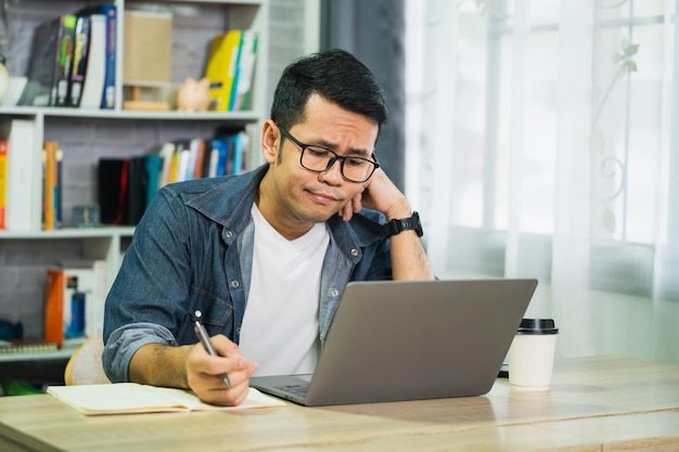 Hombre de negocios asiático usa gafas y entrega la cabeza teniendo depresión estresante momento triste mientras trabaja en una computadora portátil en casa Hombre de depresión triste serio trabajando desde casa Concepto de trabajo en cualquier lugar