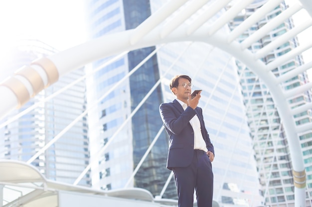 Hombre de negocios asiático usando la aplicación de teléfono móvil enviando mensajes de texto fuera del edificio de oficinas