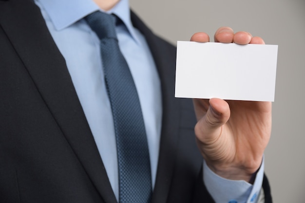 Hombre de negocios, asimiento de la mano del hombre de negocios que muestra la tarjeta de visita - primer plano sobre fondo gris. Muestre una hoja de papel en blanco. Tarjeta de visita en papel.
