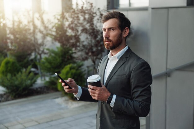 Hombre de negocios atractivo con teléfono y café parado cerca del edificio de oficinas y mirando hacia otro lado