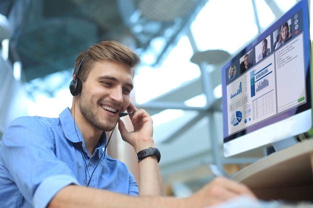 Hombre de negocios en auriculares hablando con sus colegas en videoconferencia. Equipo de negocios multiétnico que trabaja desde la oficina con ordenador PC, discutiendo el informe financiero de su empresa.