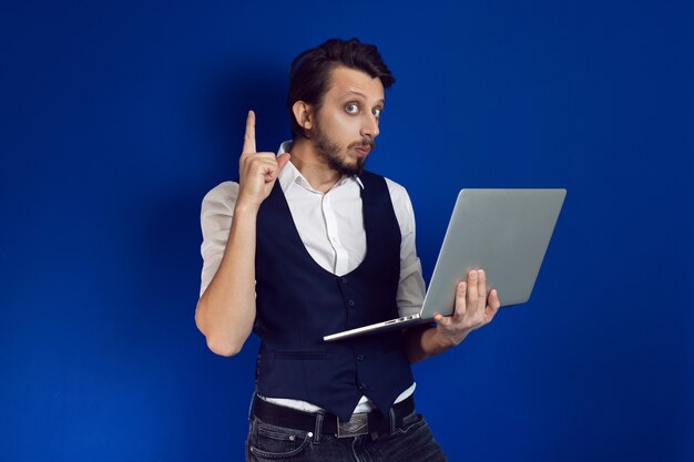 Hombre de negocios con barba en una camisa blanca y chaleco sostiene una computadora portátil en una pared azul