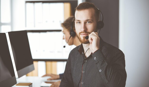 Hombre de negocios de barba roja hablando con auriculares cerca de su colega mientras está sentado en una oficina moderna. Grupo de personas diversas en el centro de llamadas. Telemarketing y atención al cliente.