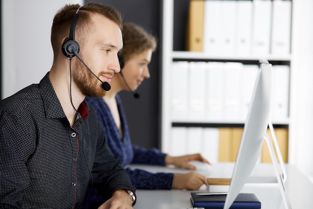 Hombre de negocios de barba roja hablando con auriculares cerca de su colega mientras está sentado en una oficina moderna. Grupo de personas diversas en el centro de llamadas. Telemarketing y atención al cliente.