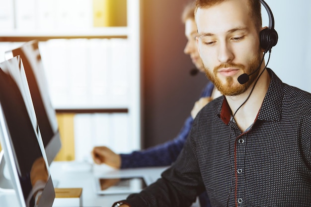 Hombre de negocios de barba roja hablando con auriculares cerca de su colega mientras está sentado en una oficina moderna y soleada. Grupo de personas diversas en el centro de llamadas.