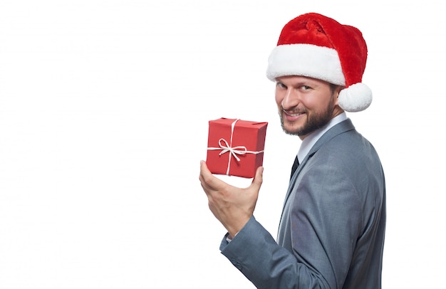 Hombre de negocios barbudo alegre en el sombrero de la Navidad que sonríe sobre el hombro que guarda el pequeño regalo de la Navidad.