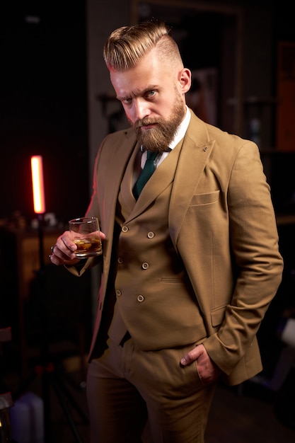 Hombre de negocios barbudo caucásico joven bebiendo un vaso de whisky en interiores en una habitación oscura