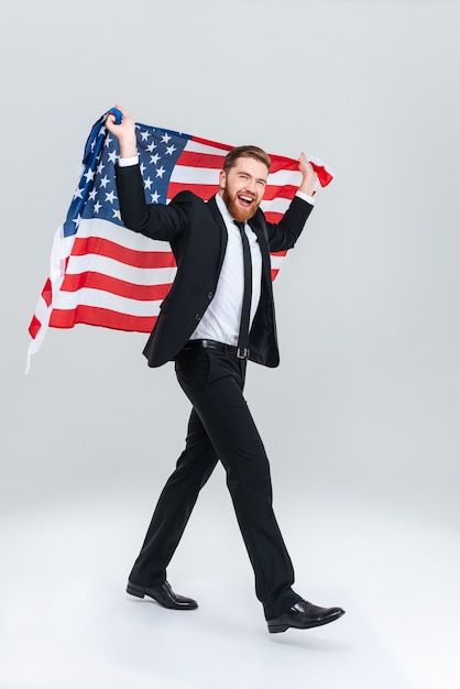 Hombre de negocios barbudo feliz de longitud completa en traje negro sosteniendo la bandera de Estados Unidos desde la parte posterior. Vista lateral. Fondo gris aislado