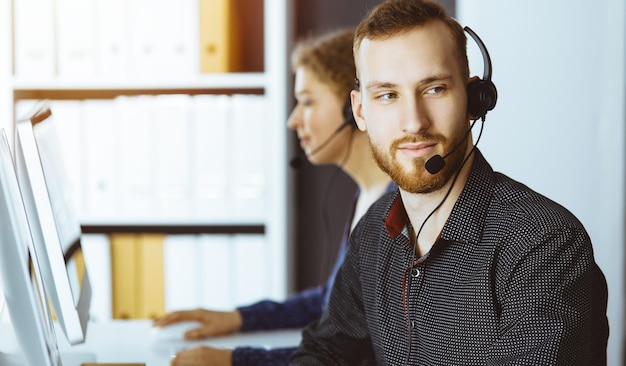 Hombre de negocios barbudo hablando por auriculares cerca de su colega mientras está sentado en una oficina moderna y soleada Grupo de personas diversas en el centro de llamadas