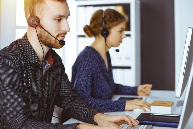 Hombre de negocios barbudo hablando por auriculares cerca de su colega mientras está sentado en una oficina moderna y soleada Grupo de personas diversas en el centro de llamadas