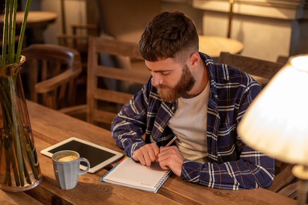 Hombre de negocios barbudo joven se sienta en casa de café en la mesa y escribe en el cuaderno cerca de la computadora de la tableta de mentiras ...