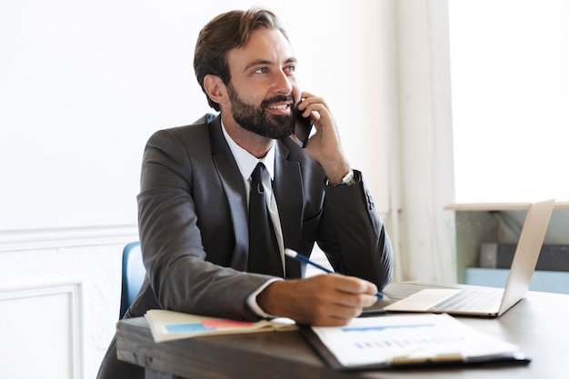 Hombre de negocios barbudo optimista complacido hermoso que se sienta en la oficina usando la computadora portátil que trabaja en interiores hablando por teléfono móvil.