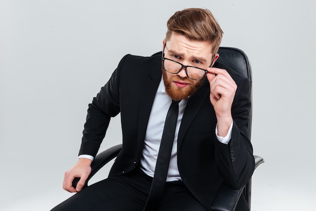 Hombre de negocios barbudo serio con gafas mirando a cámara y sentado en un sillón. Fondo gris aislado