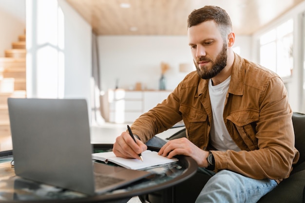 Hombre de negocios barbudo tomando notas usando una laptop sentada en casa