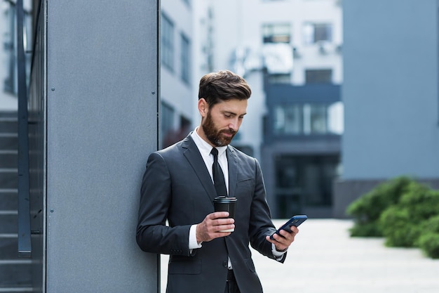 Hombre de negocios barbudo en traje formal en descanso con teléfono inteligente de uso de teléfono móvil. Hombre de negocios de pie afuera en el fondo de la calle de la ciudad urbana moderna con una taza de café en el centro al aire libre. copia espacio