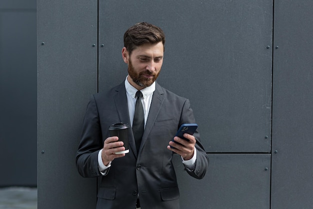 Hombre de negocios barbudo en traje formal en descanso con teléfono inteligente de uso de teléfono móvil. Hombre de negocios de pie afuera en el fondo de la calle de la ciudad urbana moderna con una taza de café en el centro al aire libre. copia espacio