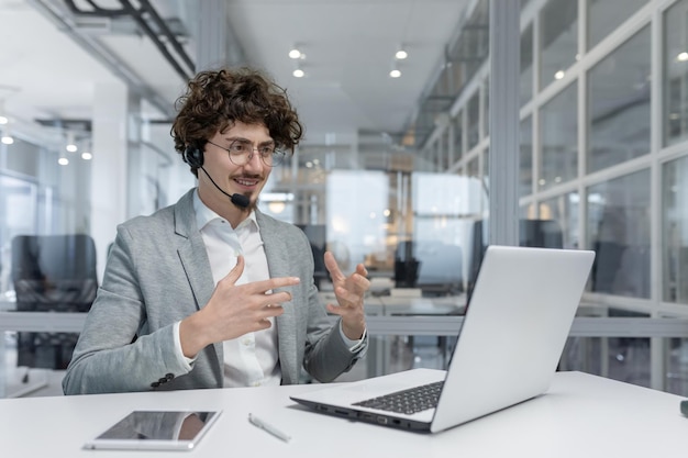 Un hombre de negocios de cabello rizado conversa en auriculares mientras trabaja en una computadora portátil en un entorno de oficina moderno