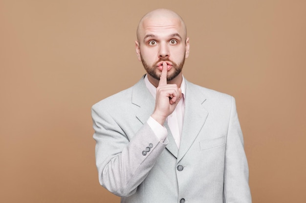 Hombre de negocios calvo en traje gris claro clásico de pie y mirando a cámara con signo de silencio