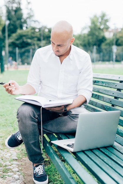 Hombre de negocios en camisa blanca que trabaja con la computadora portátil que se sienta en un banco en un parque