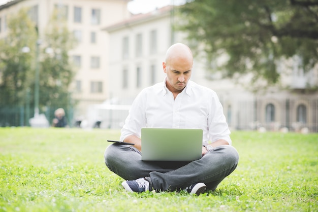 Foto hombre de negocios en camisa blanca que trabaja con la computadora portátil que se sienta en la hierba en un parque