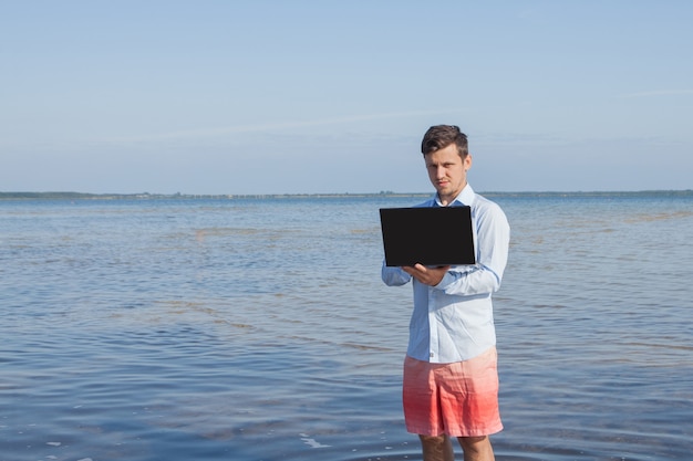 Hombre de negocios con camisa elegante y bañador con un portátil en el mar