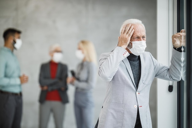 Un hombre de negocios cansado con máscara protectora parado en una oficina moderna y mirando por la ventana durante el virus de la corona o la pandemia de COVID-19.